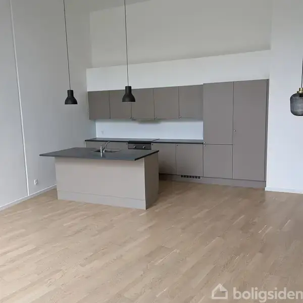 Tomt rum med store vinduer, der giver udsigt til en grå gårdhave med græsarealer og høje bygninger. Lyse trægulve og hvide vægge.