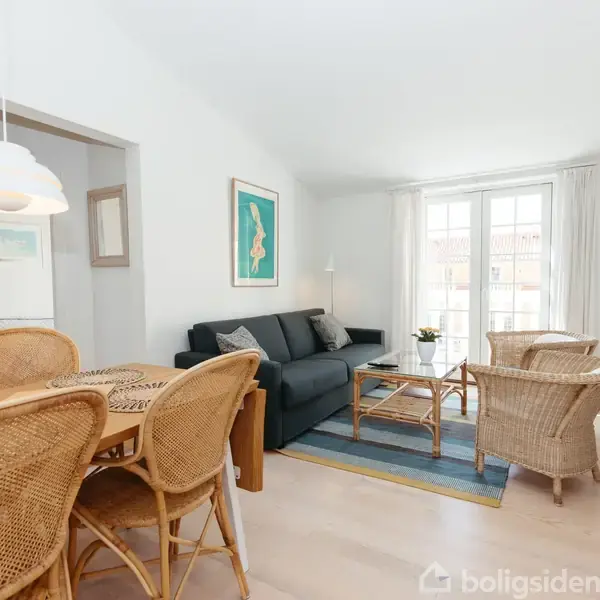 En stue med en mørk sofa står ved siden af et sofabord. To kurvestole omkranser bordet. Et spisebord med kurvestole ses til venstre. Væggene er hvide, og et maleri hænger over sofaen. En stor vinduesparti slipper lys ind til det lyse rum. En fladskærm hænger på væggen.