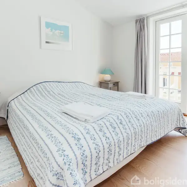 En bred seng med hvidt og blåt stribet sengetæppe ligger i et lyst soveværelse. To natborde med lamper står ved siden. En dør med glasruder fører ud til en balkon.