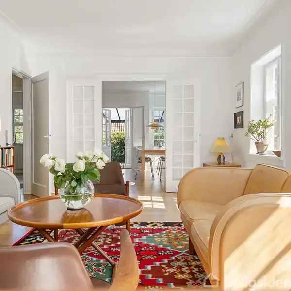 En stue med lyse vægge viser en sofagruppe omkring et rundt træbord med en vase med blomster. Farverig tæppe pryder gulvet. Sollys strømmer ind gennem vinduet.