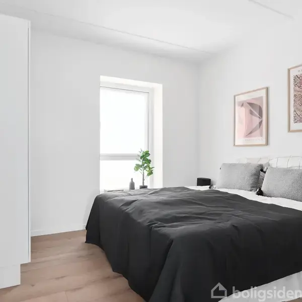 Seng med sort sengetøj står i et lyst soveværelse. Væggene er hvide, og der hænger to billeder. En vindueskarm med en plante. Trægulv og hvid garderobe ses også.