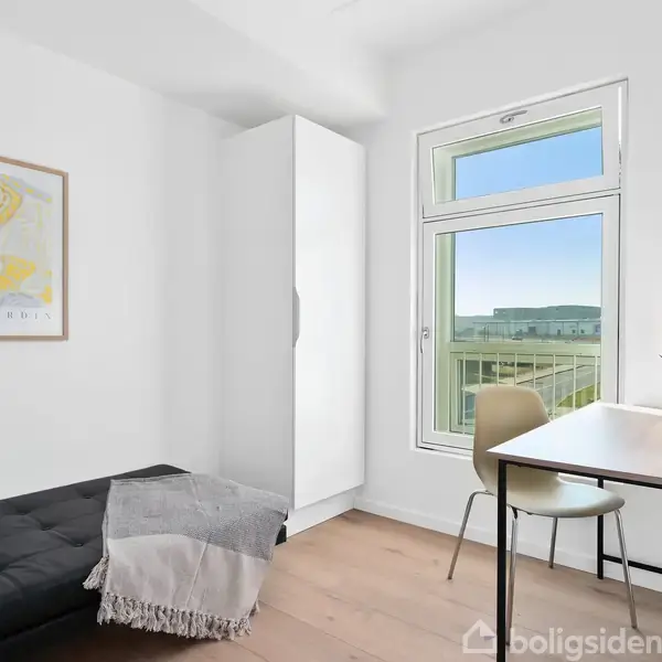 Sort sofa med puder og tæppe står ved siden af et hvidt skab. Til højre ses et lille skrivebord med stol og lampe, foran et stort vindue.