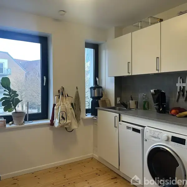 Et køkken med hvide skabe og mørke bordplader har en vaskemaskine og køkkenudstyr på bordet. To store vinduer giver udsigt til en bygning og træer udenfor.