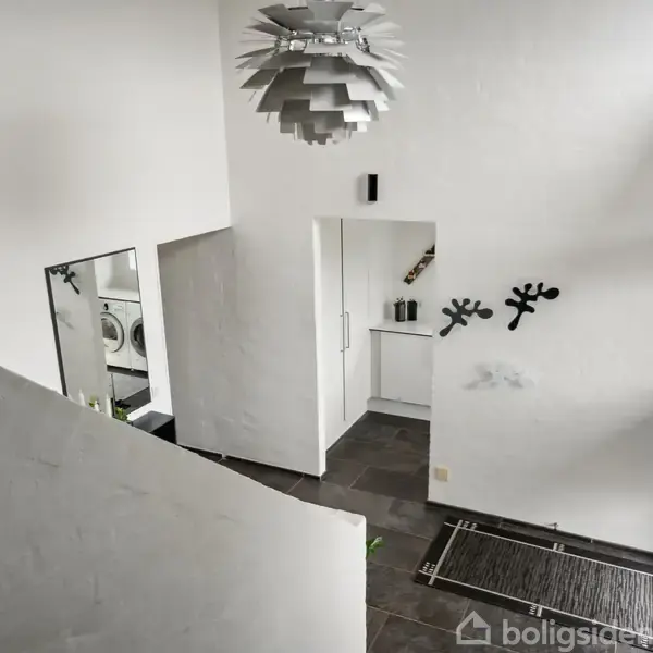 En moderne entré med hvidmalede vægge og mørke gulvfliser. En unik, geometrisk loftslampe hænger centralt. Vaskemaskine og tørretumbler ses i baggrunden gennem en døråbning.
