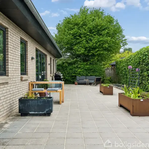 En bred flisebelagt terrasse støder op til et murstenshus med store vinduer. Terrassen har blomsterkasser og et træbord, omgivet af grøn frodig vegetation og grussti i baggrunden.