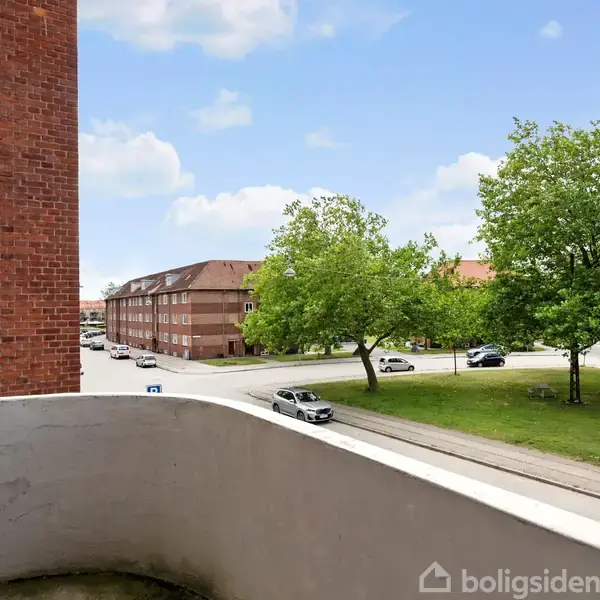 En balkon med udsigt til en vej og grønne træer. En rød murstensbygning til venstre, biler kører på vejen, og himlen er blå med skyer.