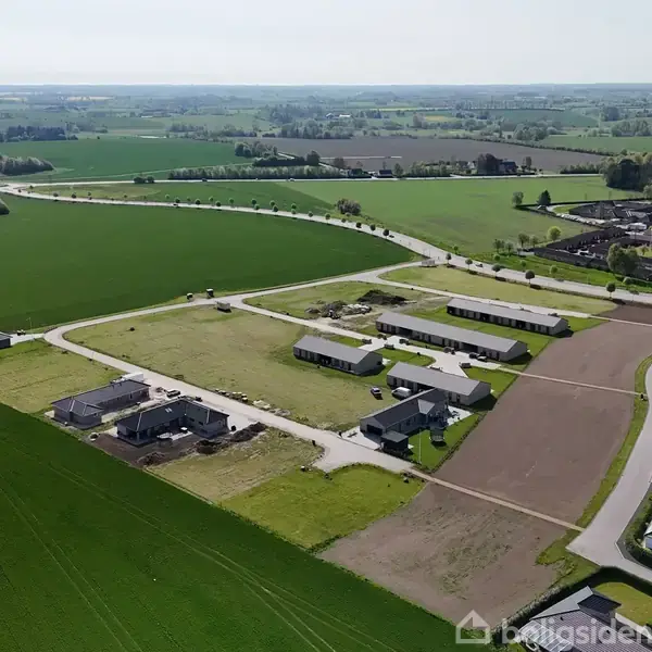 Bebyggelse set fra luften omgivet af grønne marker. Flere huse er placeret langs snoede veje i et åbent landskab, hvilket skaber en landlig atmosfære.