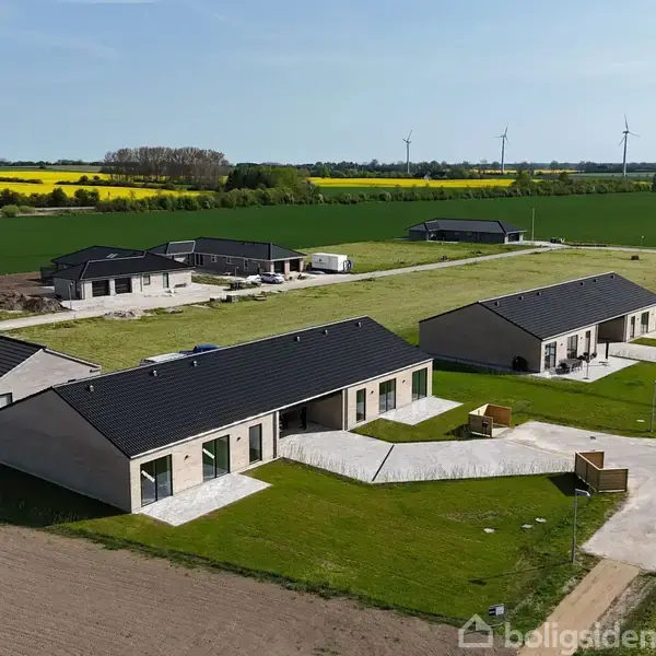 En række moderne enfamilieshuse står stille på en grøn græsplæne, omgivet af åbne marker. I baggrunden er der vindmøller, og markerne er delvist gule fra afgrøder.