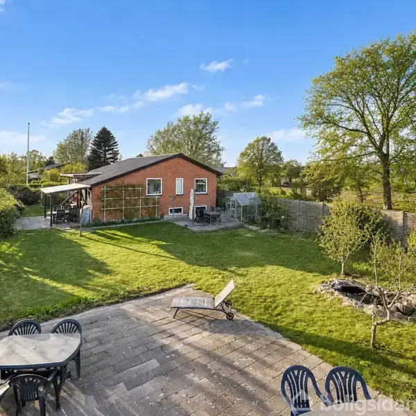 Rødt murstenshus står i en grøn have omkranset af træer. Forrest ses en terrasse med havemøbler og en solstol. Græsplæne med et mindre bålsted til højre.