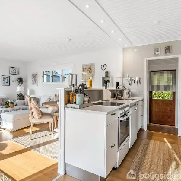 Køkkenet har hvide skabe og apparater, giver ud til en lys stue med sofaer og træmøbler. Store vinduer lader naturligt lys strømme ind i rummet.