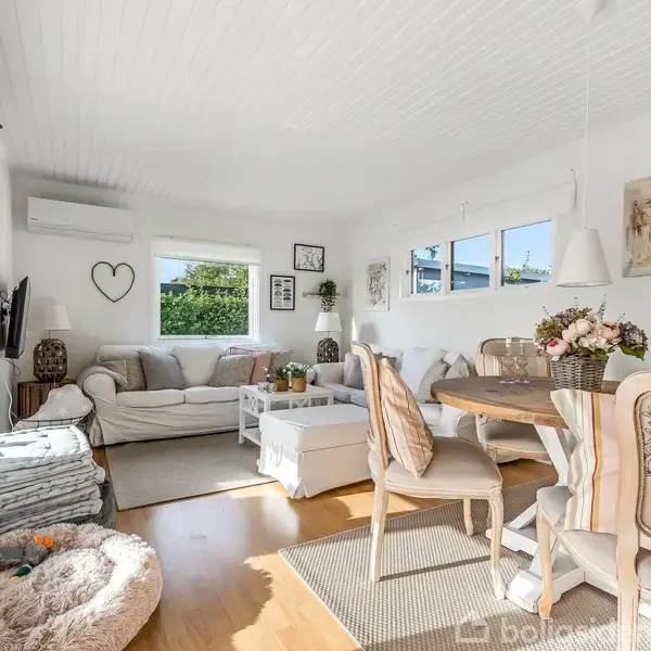 En lys stue med hvide vægge og loft. To sofaer og et sofabord står ved vinduerne. Et rundt spisebord med stole er dekoreret med blomster.