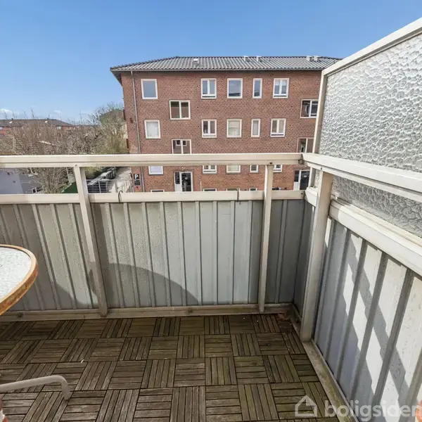 En lille balkon med trægulv og hvidt rækværk har et rundt bord. Baggrunden viser en fleretagers murstensbygning og en klar, blå himmel. Ingen tekst er synlig.