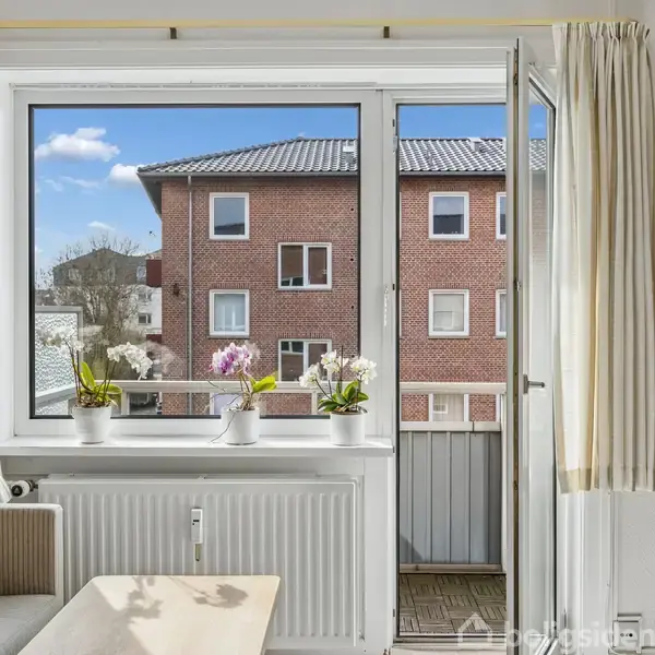 Stue med åben altandør, sofa og bord foran et stort vindue. Tre potteplanter står på radiatoren. Udsigt til rødstensbygning og blå himmel udenfor.