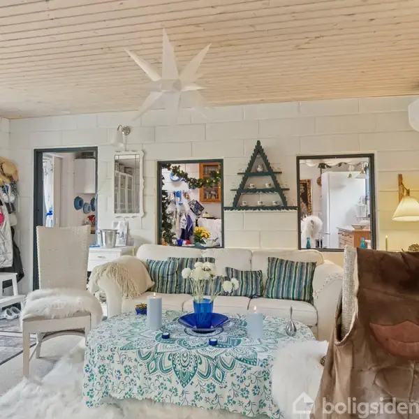 Stue med hvid sofa og farverig pude, der står foran et bord dækket med mønstret dug. Der hænger en stjerne i loftet. Vægge dekoreret med billeder og kunstværker.