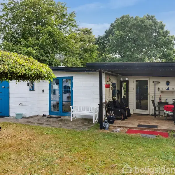 Et hvidt, enkelt hus med blå dør og vinduer. Foran er der en overdækket terrasse med møbler. Græsplænen omgives af træer og frodig bevoksning.