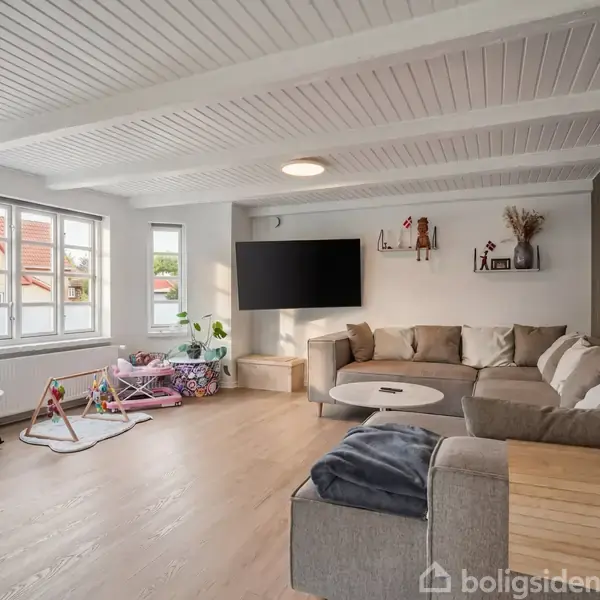 Glasdør står åben mod terrasse med sorte havemøbler og stor parasol. Omkranset af grønne buske og et hus i baggrunden. Indendørs ses et vægur på en hvid væg.