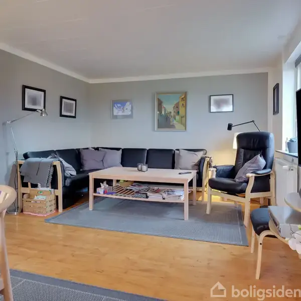 Stue med gråt sofahjørne centralt, træbord foran med magasiner og en lampe i hjørnet. Væggene er pyntet med billeder, og et stort tv står til højre.