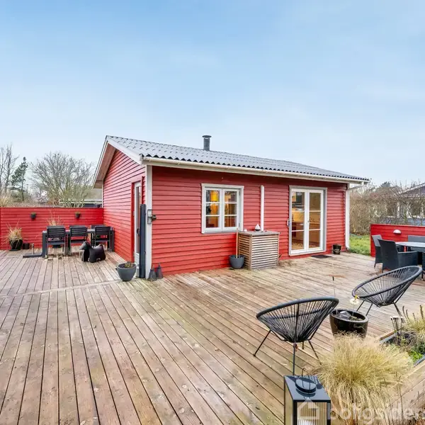 Rødt træhus med hvide vinduer står i midten af en stor træterrasse. Rundt om er der havemøbler og planter, og græsplæne omgiver terrassen. Det er dagslys og himlen er klar.