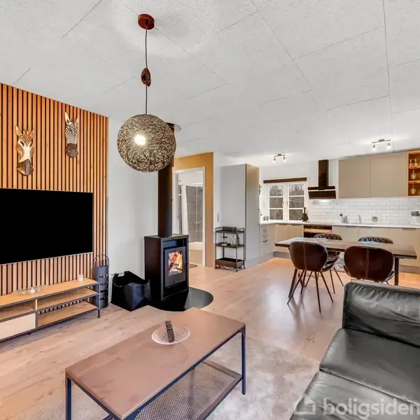 TV hænger på en væg med træpaneler, ved siden af en sort brændeovn i en moderne stue med lyst trægulv. Køkkenet er synligt i baggrunden.