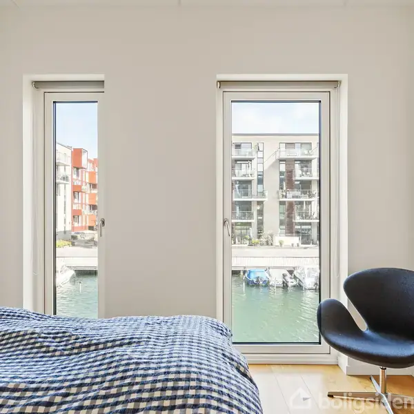 Senge med blåternet sengetøj står ved siden af to gulv-til-loft-vinduer med udsigt over en kanal. En sort stol er placeret nær væggen, som har kunst hængende.