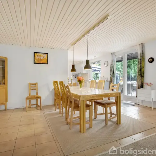 Spisebord med otte stole står i et lyst spisestueområde. Hylde og stoler ved siden af, lamper hænger over bordet. Store vinduer viser en grøn have udenfor.