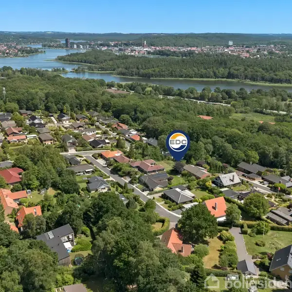 Bebyggelse med huse spredt blandt træer, set fra luften, nær en flod og sø. Et EDC-skilt markerer en bestemt ejendom i midten af billedet.