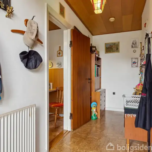 En entre med jakker hængende på væggen ved siden af en radiator. En åben dør fører til et rum med en træstol. Flere dekorationer på væggene.