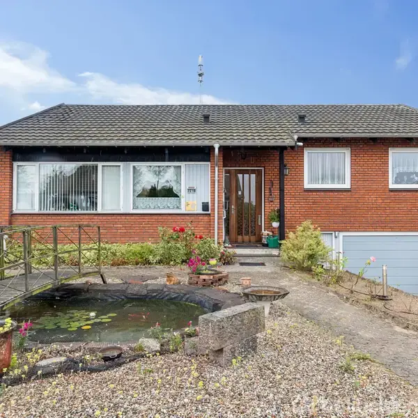 En rød murstensvilla med havebassin foran, indeholder flydende åkander. Omgivet af grus og blomsterbede. To vinduer og en hoveddør på facaden. Glimt af en garage til højre.