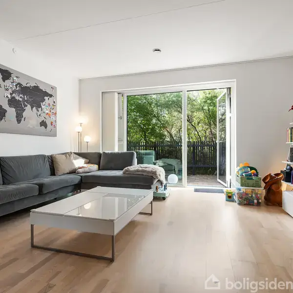 Sofa i en lys stue med trægulv, vender mod et sofabord. Skydedøre åbner ud til en have. Verdenskort på væggen, legetøj ved reolen.