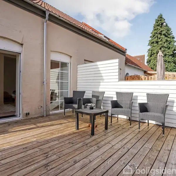 Terrasseområde med fire grå fletstole og et lille bord, placeret på trædæk foran et åbent glasdørspart til et hus. Blå himmel og have i baggrunden.