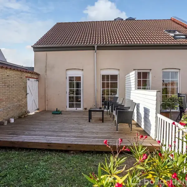 Et hus med rødt tegltag og fem hvide dobbeltvinduer. En træterrasse med havemøbler foran huset. Ved siden står en murstensbygning. Foran er en græsplæne med blomster.