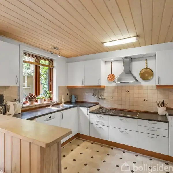 Køkken med hvide skabe og sorte bordplader har planter ved vinduet, køkkenredskaber hængende på væggen og en trælaminatloft. Fliser på væggen og et blomstermønstret gulv skaber en hyggelig atmosfære.
