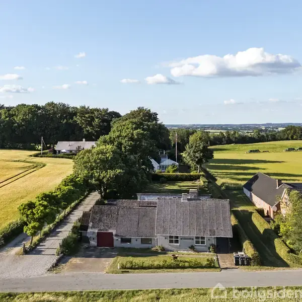 Huset står stille og omgivet af marker med en grusvej foran. Baggrunden viser skove og en blå himmel med spredte skyer.