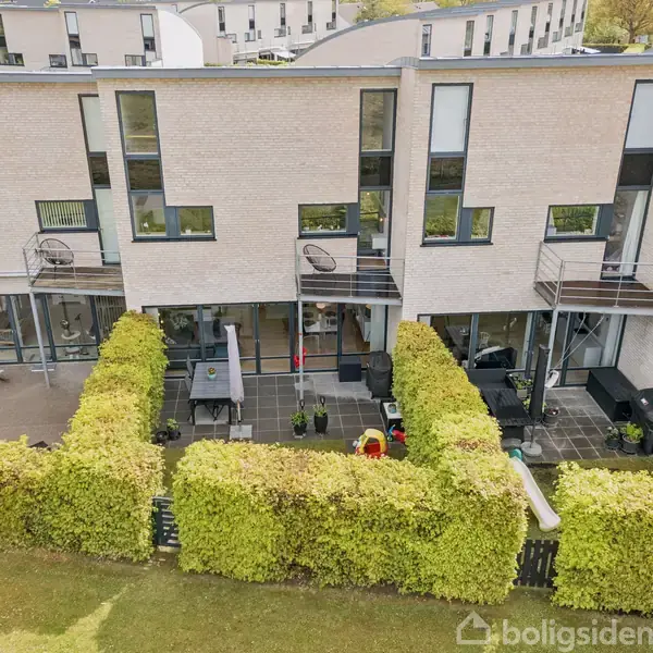 En række moderne lejligheder med altaner og terrasser i en baggård. Terrassen er omgivet af grønne hække og indeholder en grill og havemøbler.
