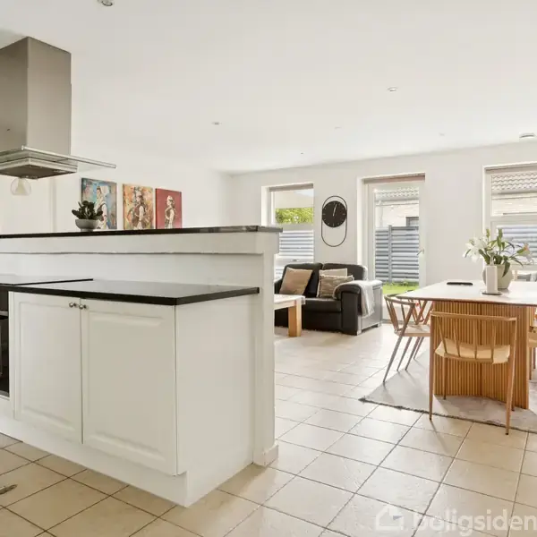 Køkken-alrum med hvidt køkkenmodul og emhætte. Spisebord med stole og planter. Sofa ved væg med kunst. Store vinduer giver lys. Flisegulv i rummeligt, moderne interiør.