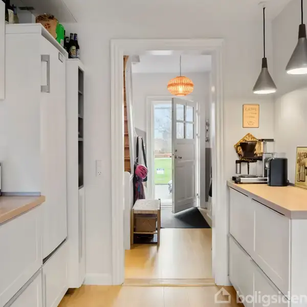 Et smalt køkken med hvide skabe og træbordplade. En døråbning fører til en lys gang med udsigt til haven udenfor. En kaffemaskine står på bordpladen.