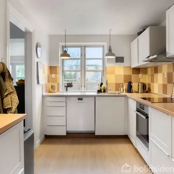 Et lyst, moderne køkken med hvide skabe og træbordplader. Der er to vinduer over vasken, og væggene har ternede fliser i beige nuancer. Køkkenudstyr ses på bordet.