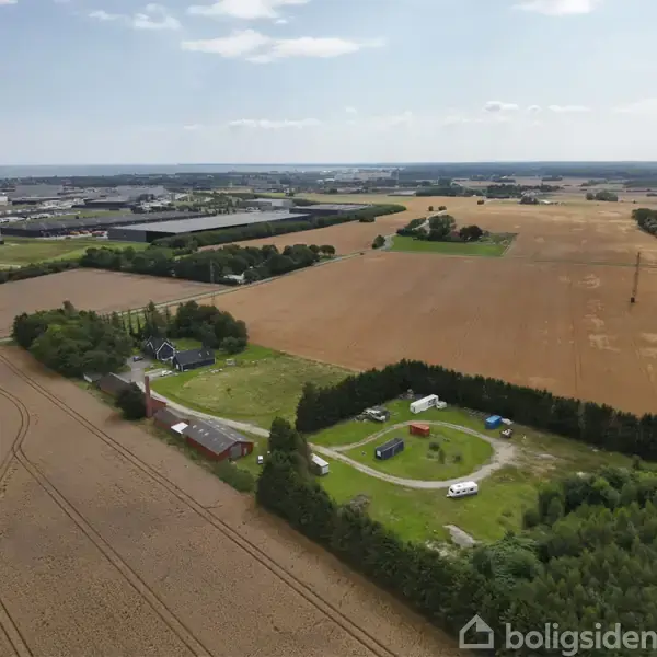 Flyfoto viser landbrug med gyldne marker. En gård omgivet af træer ligger i midten. I baggrunden ses industribygninger og flere marker, der strækker sig til horisonten.