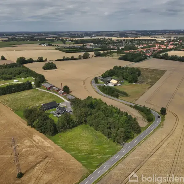 Luftfoto af landevej, der snor sig gennem marker og jordområder, omgivet af enkelte huse og skovstykker i et landligt landskab med tætliggende by i baggrunden.