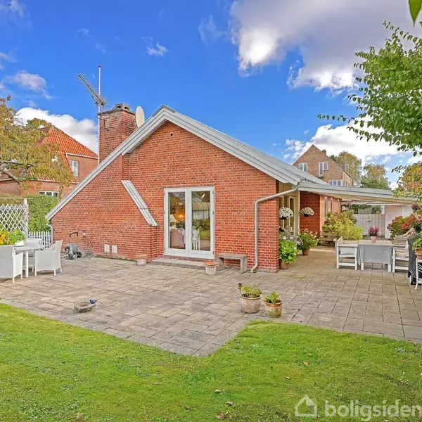 Rødt murstenshus står stille med en have omkranset af grøn vegetation. Terrasse med flisebelægning har flere havemøbler til afslapning. Klar blå himmel giver en rolig atmosfære.