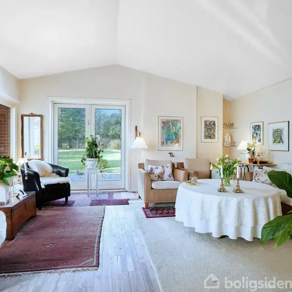 Stue med sofaer og lænestole omkring et rundt bord med dug, dekoreret med kunst på væggene; store vinduer giver udsigt til en have med græsplæne.