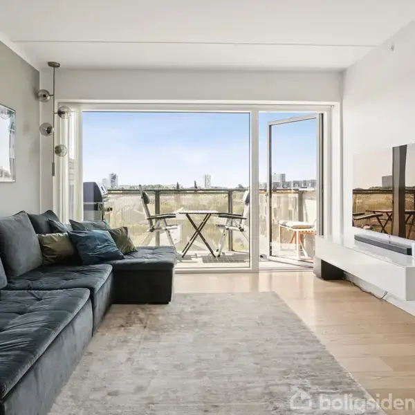 Sofa står i stuen, vendt mod et åbent vindue der fører til en balkon med et bord og stole. Et TV står på en hylde til højre.