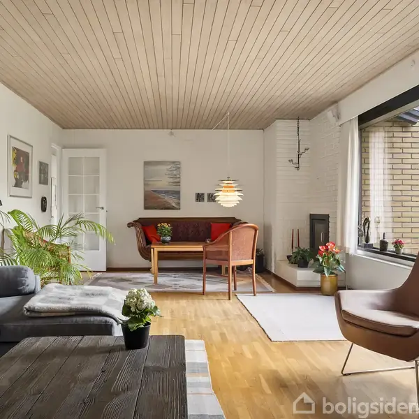 En stue med sofa og stol står på en trægulv. Et bord har en potteplante. Store vinduer giver udsigt til en have.