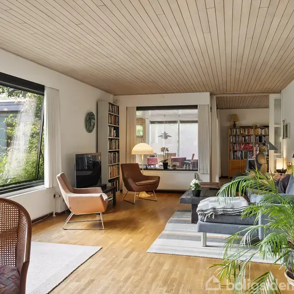 Et lyst stueområde har en grå sofa, en træstol, og et sofabord. Rum med bogreoler og store vinduer sætter en hyggelig tone, mens planter tilføjer livlighed til rummet.