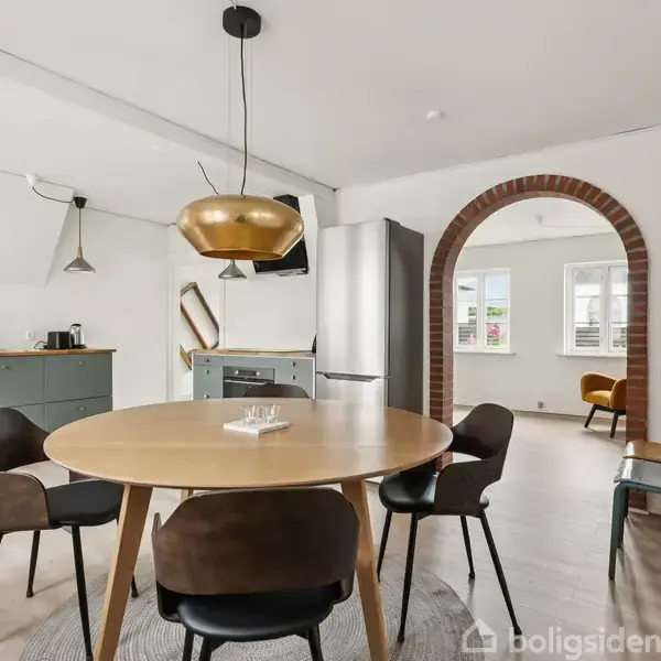 Rundt spisebord med fire stole står under en guldfarvet lampe, i et moderne køkken med hvide vægge og træmøbler. Der ses en buegang til stue med vinduer i baggrunden.