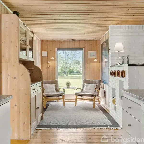 Stue med to stole foran et stort vindue, der giver udsigt til en grøn have. Køkken til venstre, hvid pejs til højre, trævægge og trægulve.