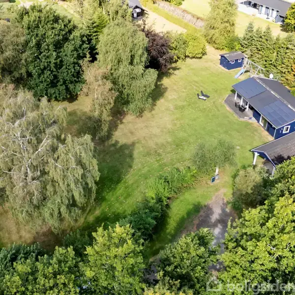 Et blåt sommerhus med en mørk tag står på en grøn græsplæne omgivet af træer og buske. En lille sti fører til huset i en skovklædt, landlig baggrund.