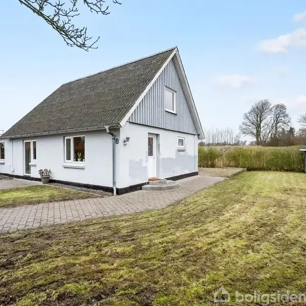 Et hvidt hus med spidst tag står stille i en velplejet have med græsplæne og flisebelagt sti. I baggrunden ses en lille grå skur, omgivet af buske og træer.