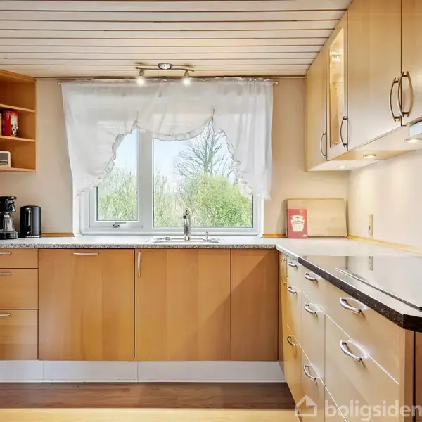 Køkken med træskabe, elkedel og kaffemaskine på bordpladen, med udsigt gennem et vindue med hvidt gardin. Hylder i hjørnet opbevarer krydderier. Grønne træer udenfor vinduet.