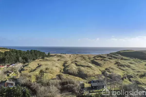 Landskab med græsdækkede klitter strækker sig mod havet i horisonten. Enkeltstående huse ligger spredt i forgrunden, omgivet af træer og buske under en klar, blå himmel.
